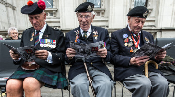 Three WWII Veterans are sat on chairs read the program for the VE Day Service ahead of the ceremony.