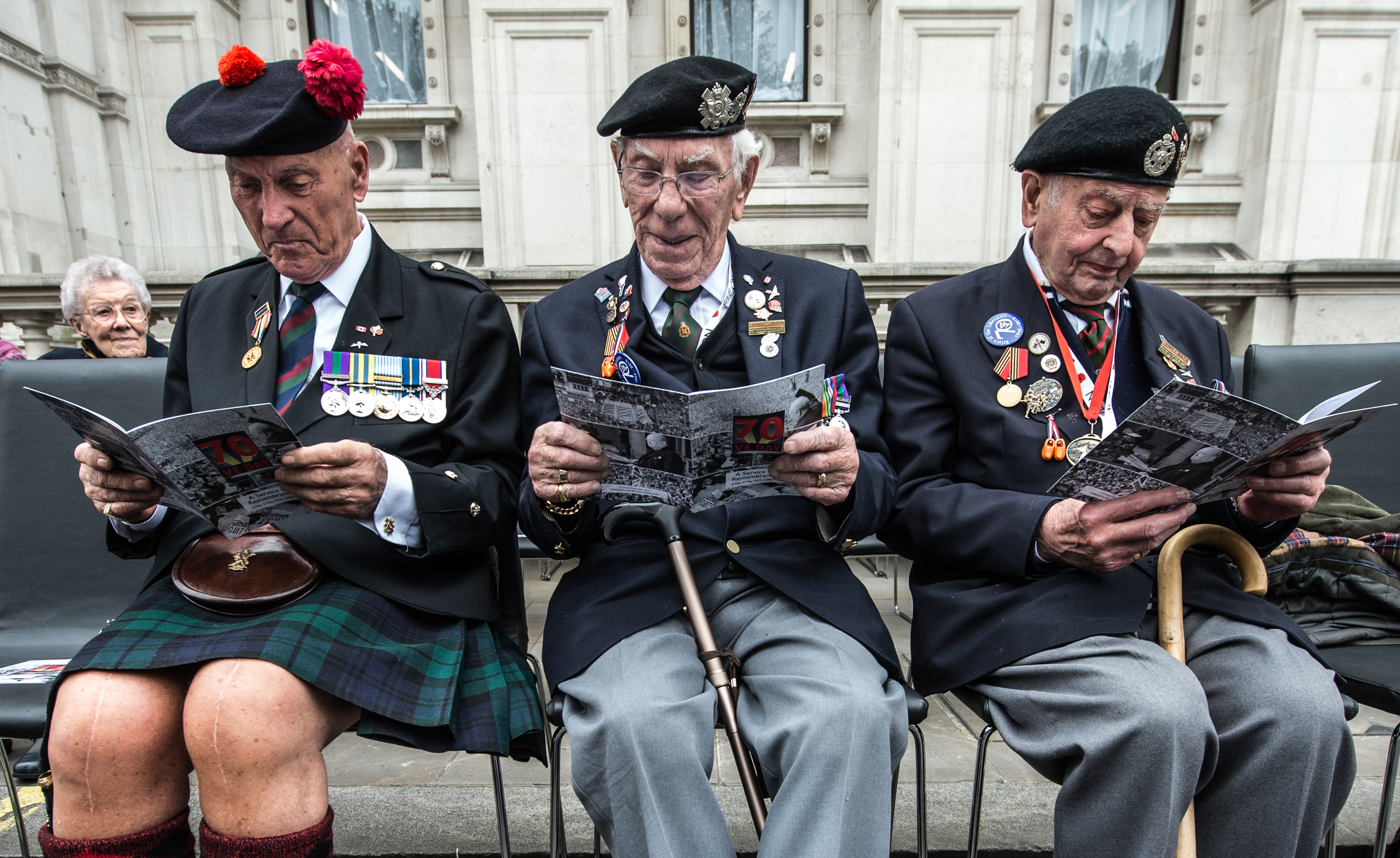Three WWII Veterans are sat on chairs read the program for the VE Day Service ahead of the ceremony.